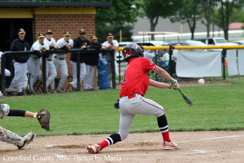 Batter in a red uniform swings at a pitch near home plate as the catcher crouches behind him and teammates watch from the dugout behind the fence.