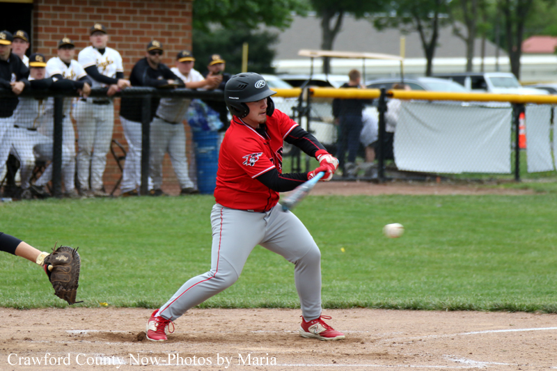 Baseball batter in a red jersey and gray pants swings at a pitched ball on a dirt infield, helmet on, with spectators behind the fence watching.