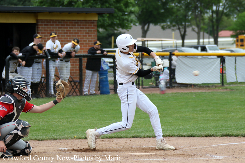 Baseball batter in a white uniform swings at a pitch near home plate as the catcher crouches behind him, with spectators and a fence in the background.