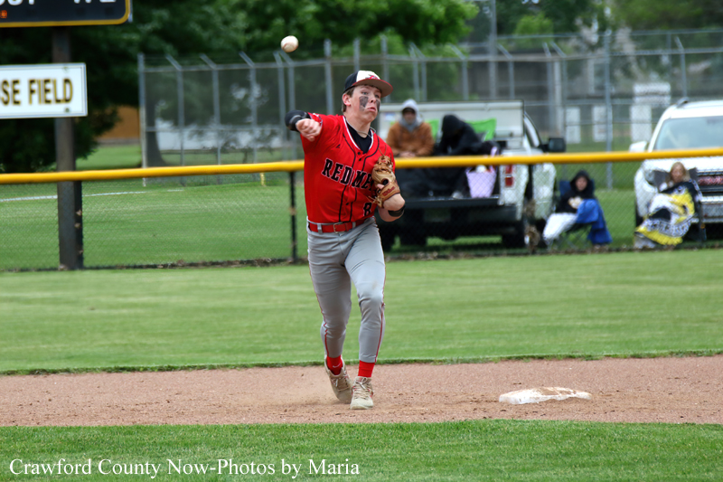 Baseball player in a red uniform throws a ball on the infield during a game, with a yellow fence and spectators in the background.