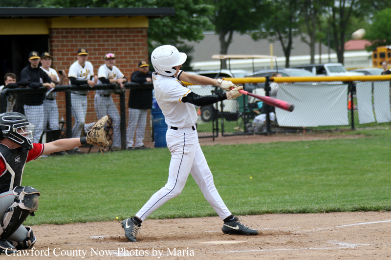 Baseball batter in a white uniform swings a pink bat during a game, with a catcher in red and black squatting behind and spectators watching from the fence.