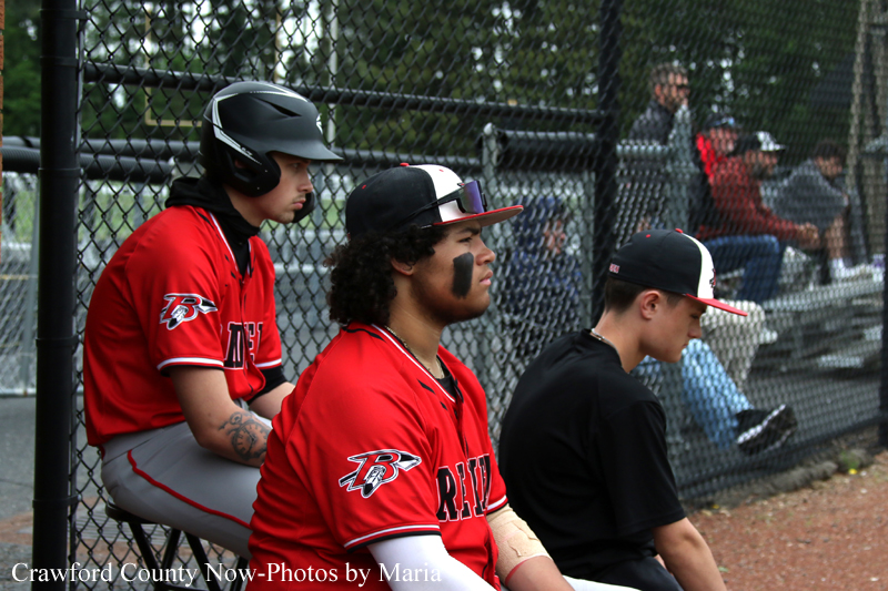 Three baseball players sit on a dugout bench beside a chain-link fence, wearing red and black uniforms; left is helmeted, center has face paint and sunglasses, right wears a black cap and shirt.