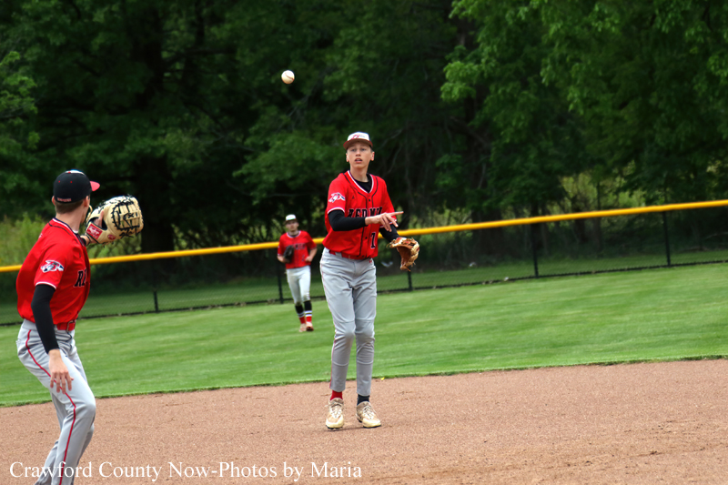 Youth baseball game: pitcher in red uniform throwing as infielders watch on a dirt infield with a green outfield and yellow foul line in the background.
