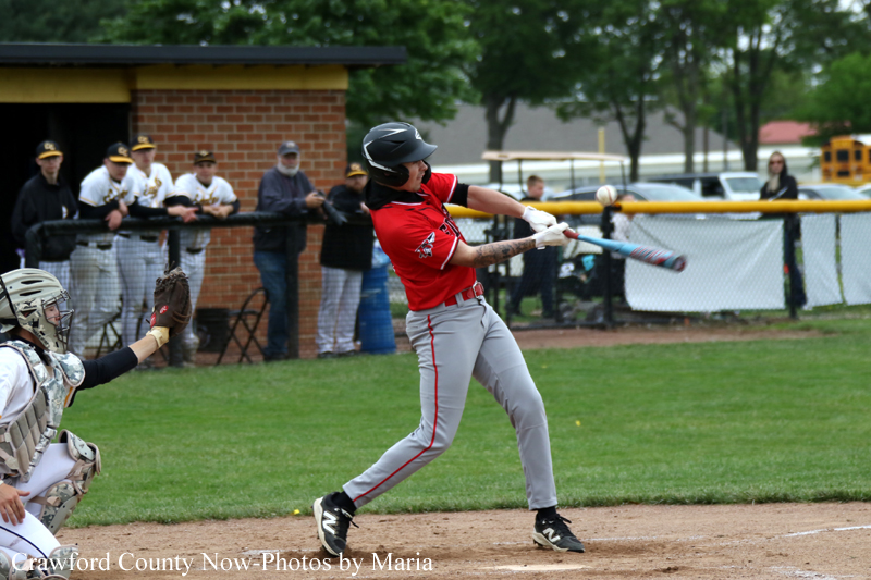 Baseball batter in a red jersey swings at a pitch as the ball approaches the bat on a dirt field with a catcher crouched nearby.