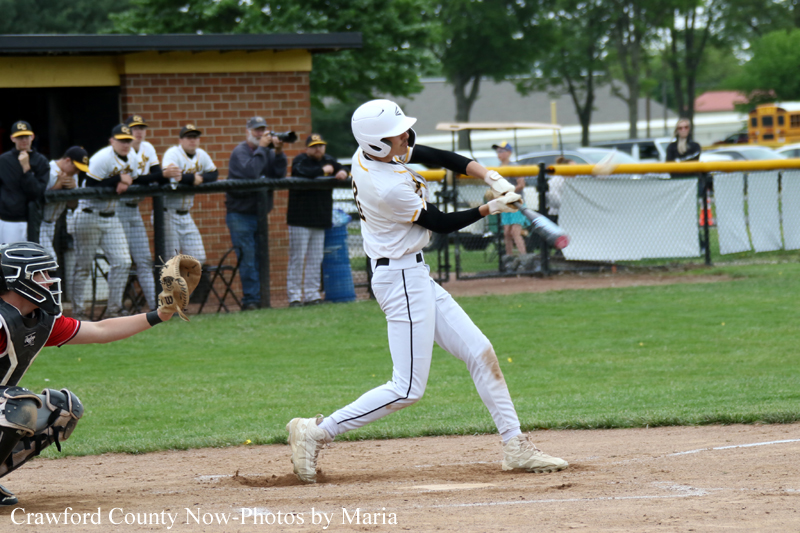 Baseball batter swings at a pitch during a game, catcher ready behind home plate and spectators in the background.