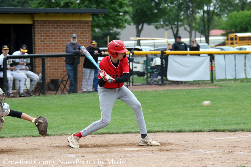 A young baseball batter in a red helmet swings at a pitched ball on a dusty infield, with a catcher in view behind him and fans in the background.