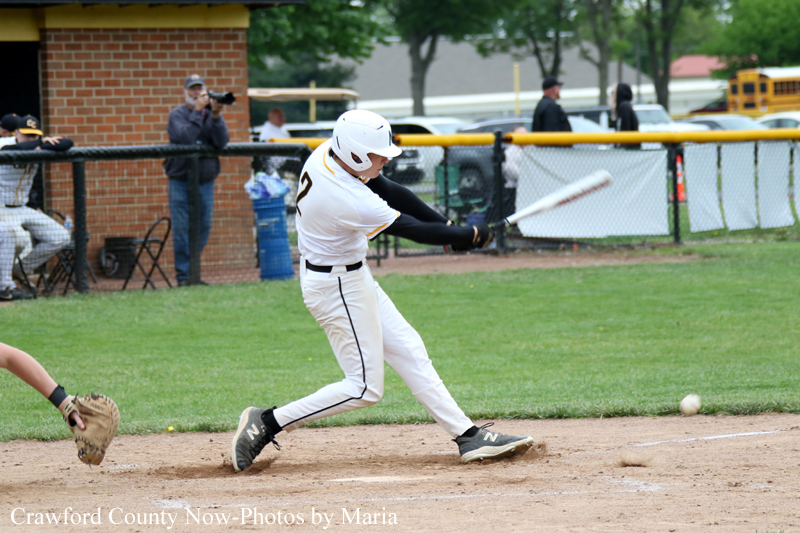 Baseball batter in white uniform swings at a pitched ball during a game on a kept grass field with a fence and onlookers in the background.