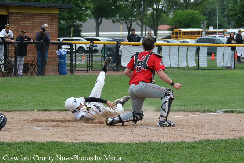 Baserunner slides into home plate as the catcher in red gear moves to tag, with fans and cars visible behind the fence in a baseball field setting.