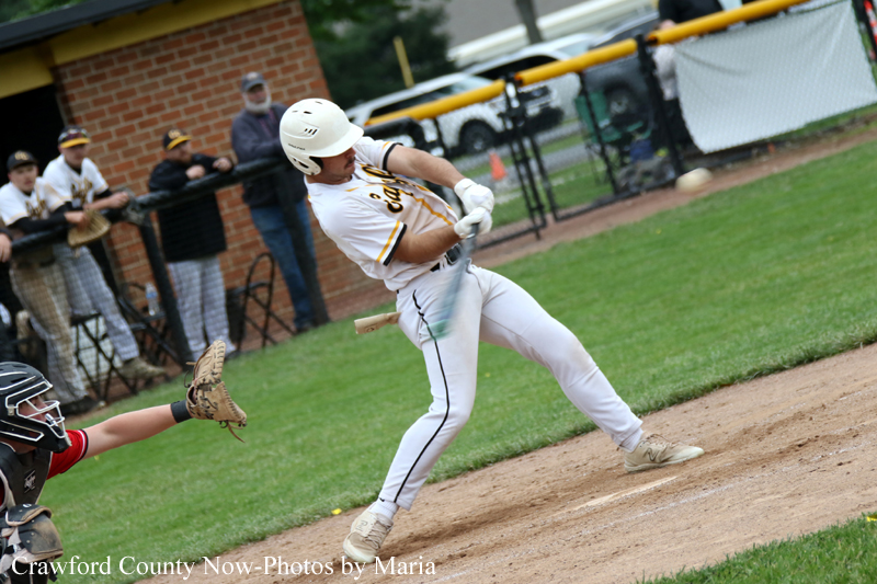 Baseball batter in a white uniform swings at a pitch near home plate, with the catcher in black gear reaching from the left and players watching in the background.