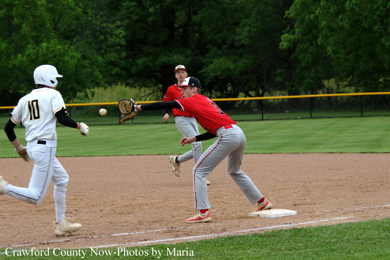 Red-team baseball fielder lunges with glove to field a ball as white-team baserunner wearing #10 advances on the dirt infield near first base.
