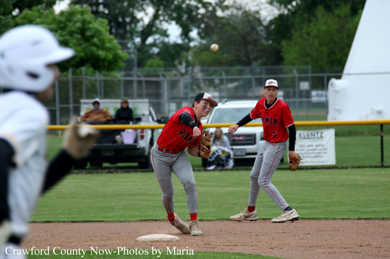 Youth baseball game scene: a fielder in red reaches for the ball near second base as a teammate watches, with a player in white in the foreground.