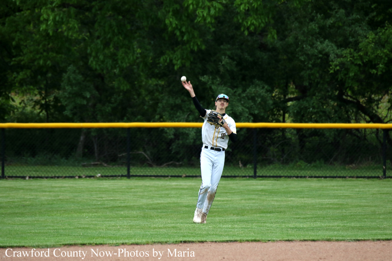 Baseball fielder in a white uniform reaches up to catch a ball in the outfield with a yellow fence and green trees in the background.