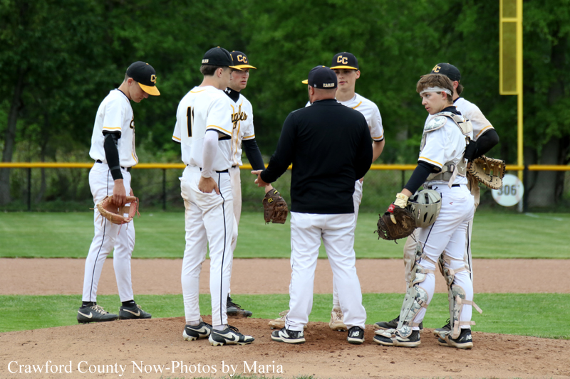 Baseball team in white uniforms huddle on the pitcher's mound as a coach talks to players, catcher in gear watches nearby on a sunny field.