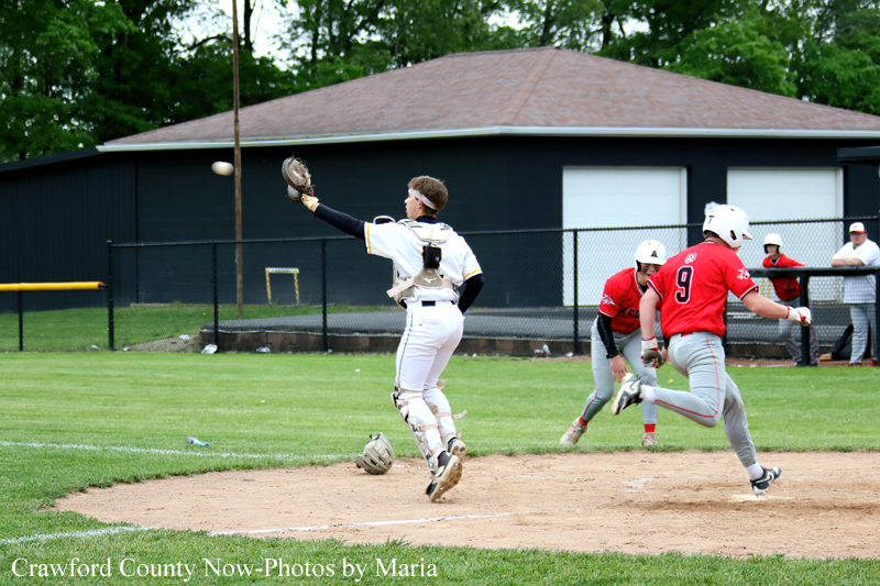 Baseball catcher in white gear stretches his glove at home plate as a baserunner in red (#9) slides in on a sunny day near a dark building.