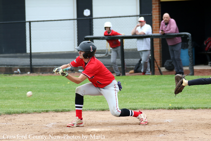 Young baseball player in a red uniform and helmet lunges to field a ground ball on the dirt infield, with a chain-link fence and spectators in the background.