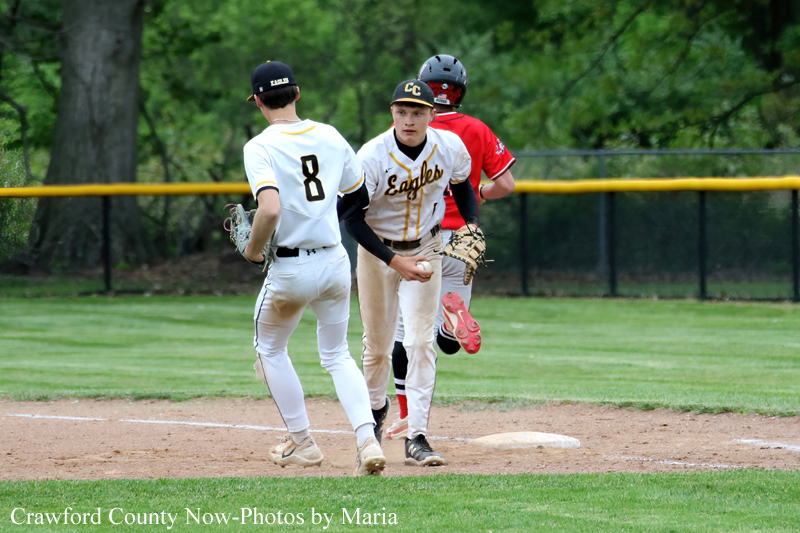 Two baseball players in white Eagles uniforms near first base; one with the ball, the other facing him, as a red-clad fielder watches behind.