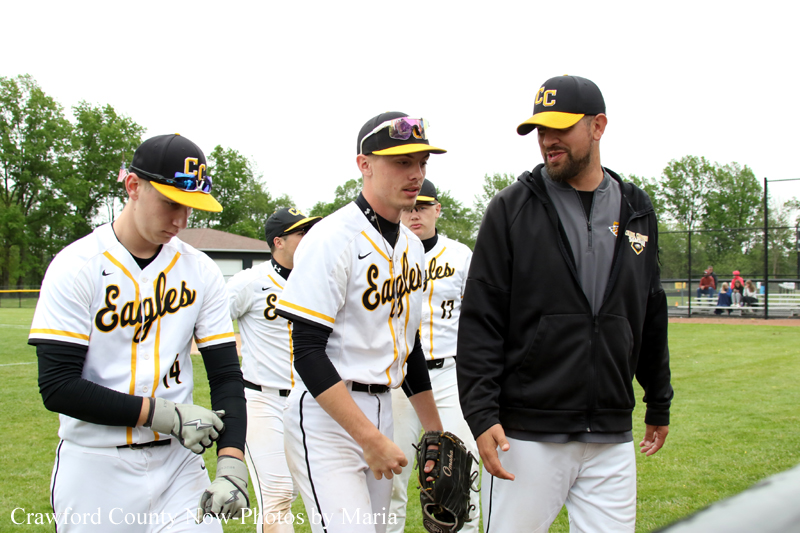 Baseball players in white and yellow Eagles uniforms walk with a coach on a grassy field after a game.