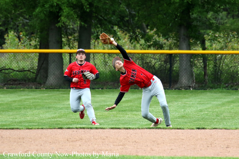 Two young baseball players in red and gray uniforms on a grass field, one leaping with his glove to catch a ball while the other moves nearby in a park-like outfield with a yellow fence behind them.