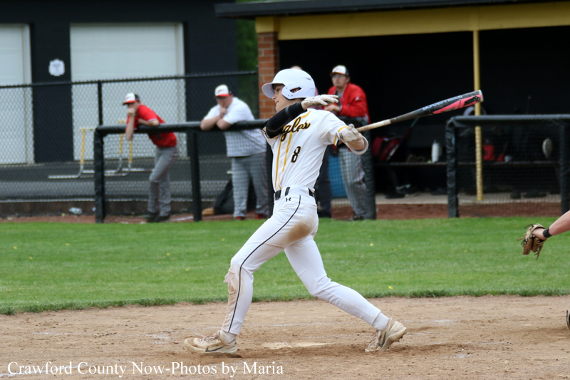 Baseball batter in a white uniform swinging a bat on a dirt infield, with teammates and a dugout in the background.