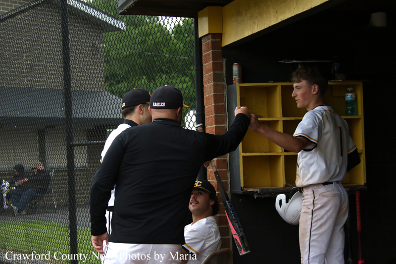 Baseball players in the dugout share a fist bump between a coach in black and a young player in white, near a yellow storage shelf.