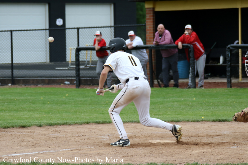 Baseball player in a white uniform (#11) runs on the dirt infield as a ball sails nearby, with spectators behind the fence.