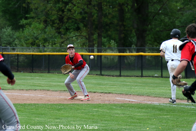 Baseball game in progress: a red-jersey fielder crouches with glove ready as a ball sails toward him, white-uniform runner nearby and outfield in the background.
