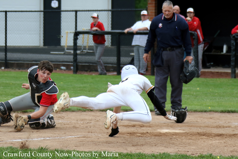 Baseball action: a white-uniformed runner slides into home plate as the catcher in black gear lunges for the tag; an umpire and teammates watch from the background.