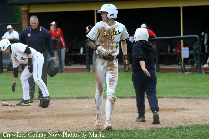 Baseball game scene: a player in a white and gold uniform and helmet walks on the dirt infield, muddy from play, while teammates and an umpire gather nearby.