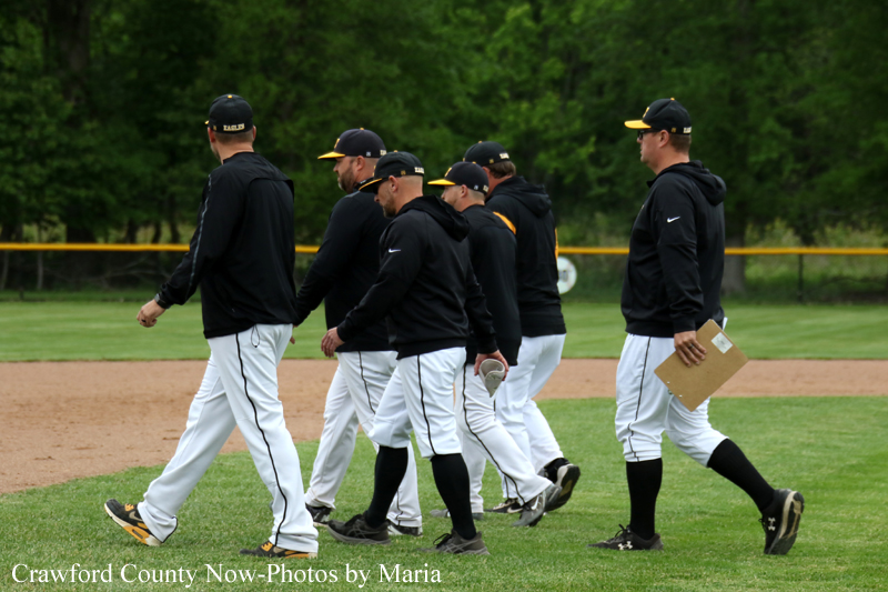 A group of baseball players in black jackets and white pants walk in a line across the infield, one carrying a glove and another a clipboard.