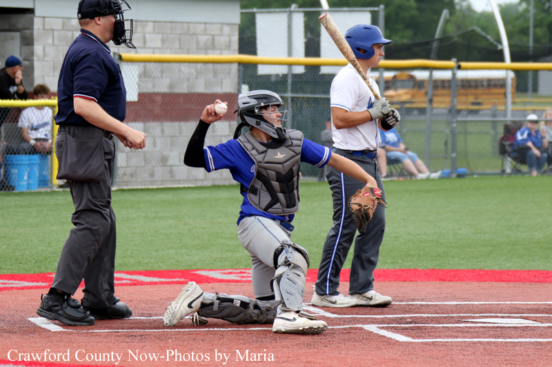 Baseball catcher kneeling at home plate in blue gear, batter in white shirt with bat, umpire nearby.