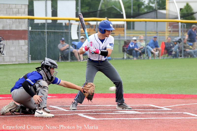 Baseball batter in a white uniform and blue helmet swings at a pitched ball, with a catcher in blue crouched behind home plate and spectators watching from the fence behind them.