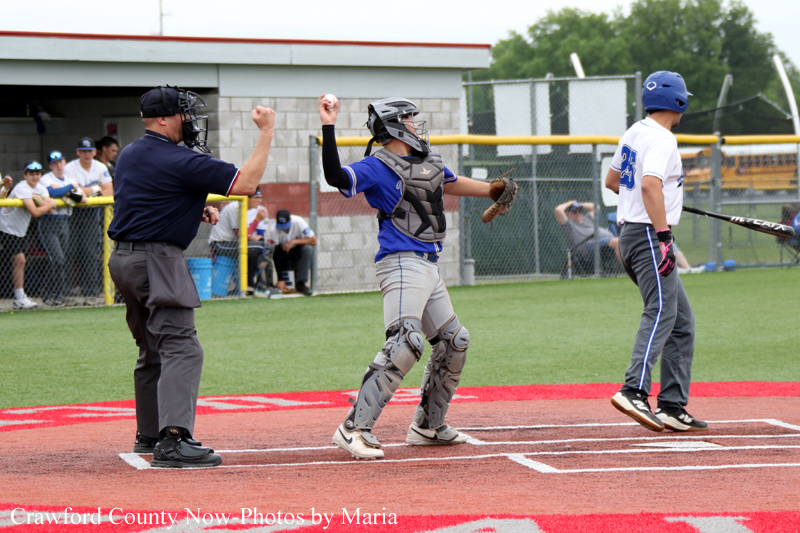 Baseball catcher in blue uniform raises arms in celebration at home plate as umpire also raises a fist and a batter walks away nearby.