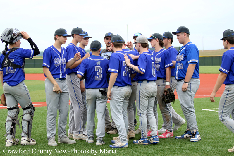 Blue-uniformed baseball team huddles on a field, listening to a coach during a pre-game talk.