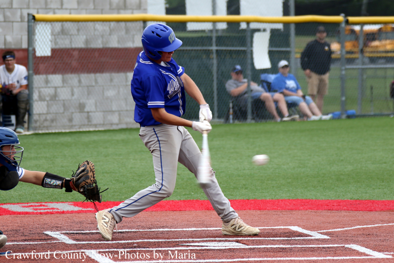 Baseball batter in a blue uniform swings at a pitched ball on a red clay field with a catcher in the foreground.
