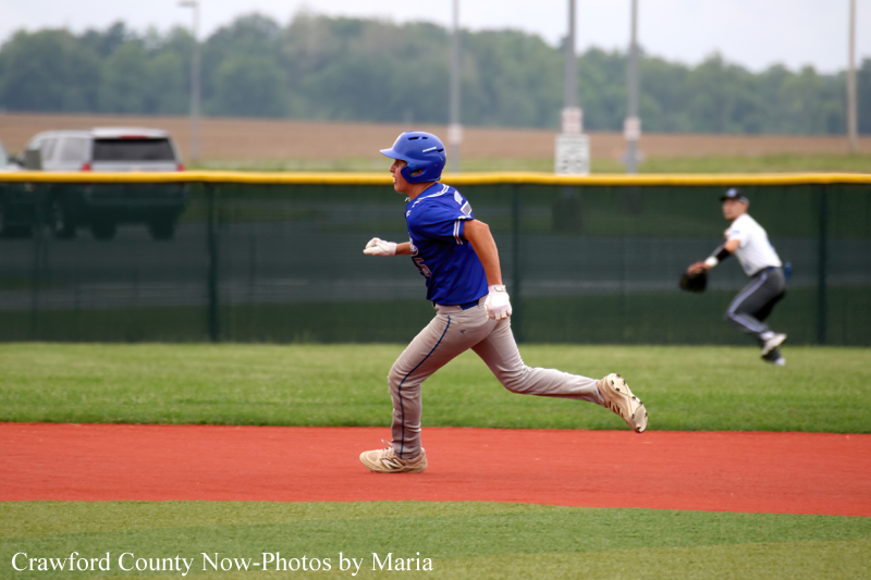 Baseball base runner in blue uniform sprinting on red dirt of the infield with a helmet and glove on his left hand at a sunny field.
