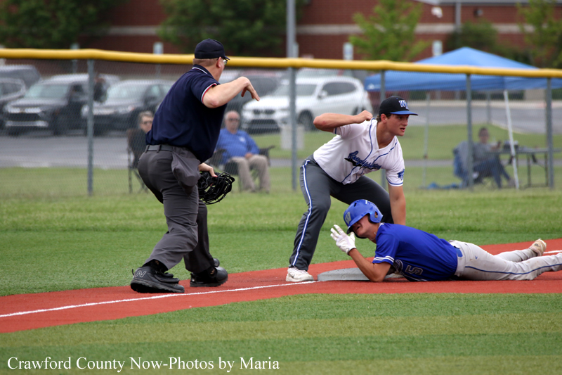 Baseball player in blue slides into a dirt base as a defender in white reaches to tag; umpire watches closely behind them.