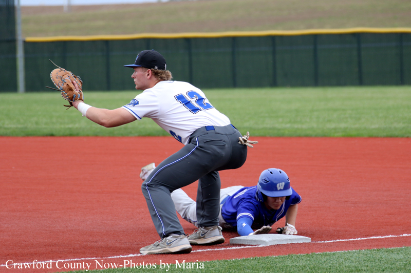 Baseball infielder in a white jersey (#12) reaches with his glove to catch a ball as a blue-clad runner slides into the base on the red clay infield.