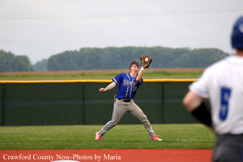 Baseball fielder in blue uniform reaches up with his glove to catch a ball on the outfield, foreground player in white blurred.