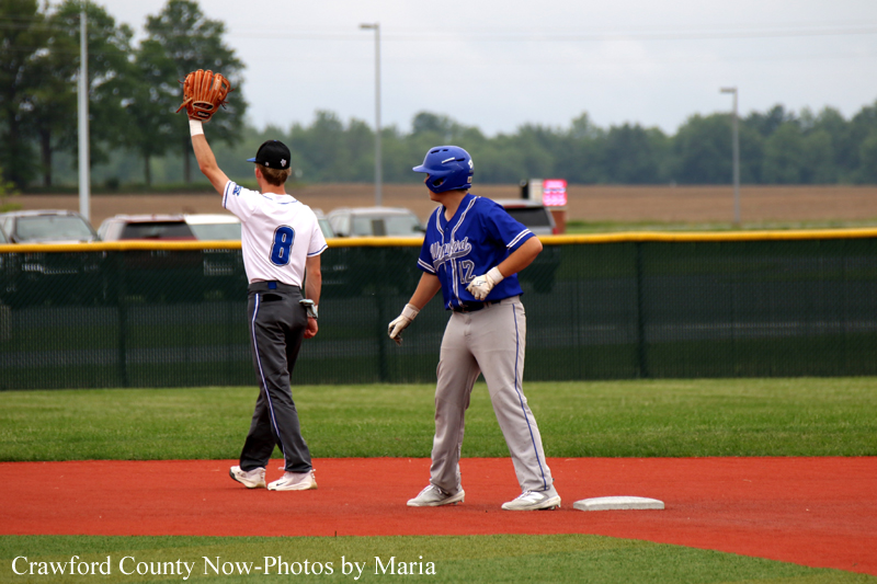 Two young baseball players stand on a dirt infield near second base: one in a white jersey with #8 raises his glove, while the other in a blue uniform stands nearby on base.
