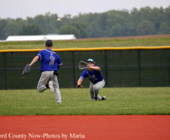 Two young baseball players in blue uniforms on a grassy field; one pitches while the other dives to catch a ball near the outfield fence.