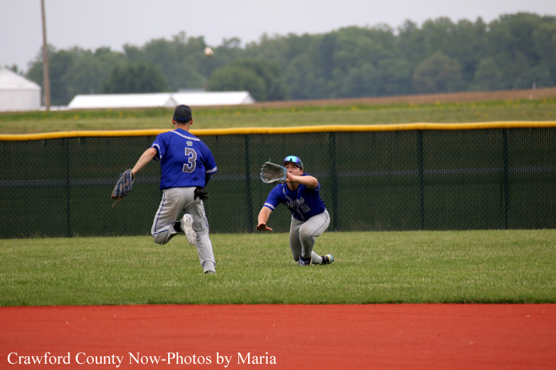 Two young baseball players in blue uniforms on a grassy field; one pitches while the other dives to catch a ball near the outfield fence.