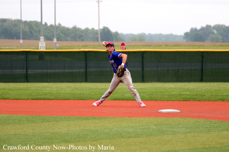 Baseball player in a blue uniform throwing a ball on the dirt infield with green outfield behind a fence.