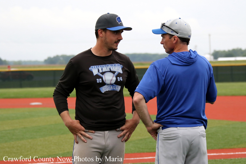 Two men in baseball caps stand on the infield, hands on hips, chatting during practice on a sunny day.