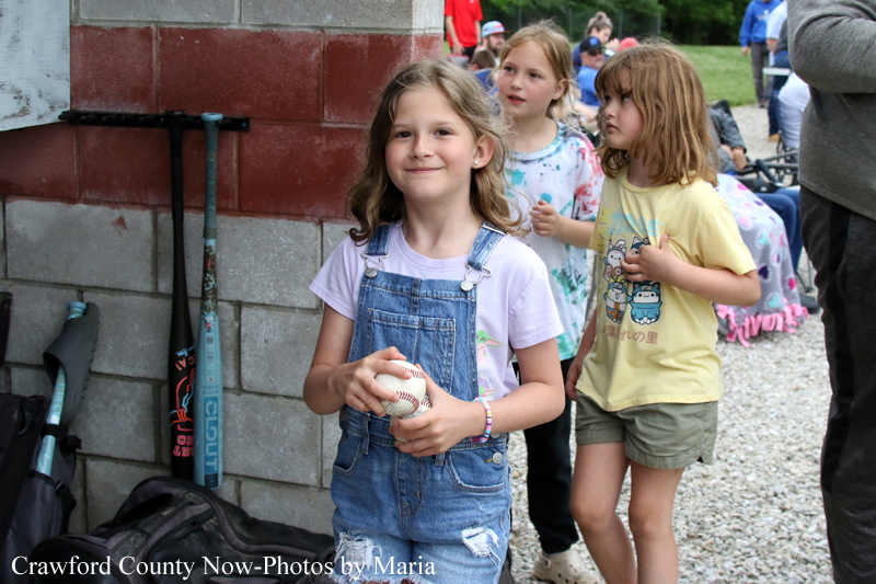 Girl in denim overalls and a light purple shirt holds a baseball, smiling near a brick wall at an outdoor event.
