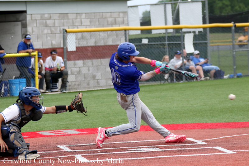 A baseball batter in a blue uniform and pink cleats swings at a pitch during a youth game, with the catcher and spectators in the background.