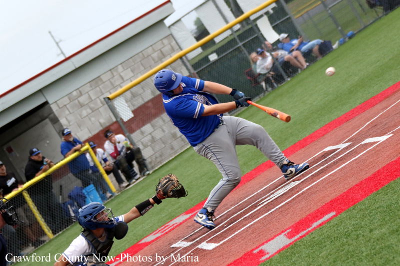 Baseball batter in blue swings at a pitch near home plate as the ball sails in front of a catcher in black gear in a fenced field stadium.