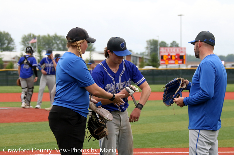 Softball players in blue uniforms on a field as a coach hands equipment to a teammate; scoreboard in the background.