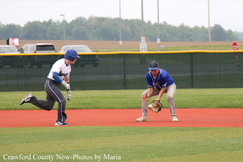 Two baseball players on a red-dirt infield: a baserunner in white and blue moves toward a blue-uniform fielder crouched with a glove.