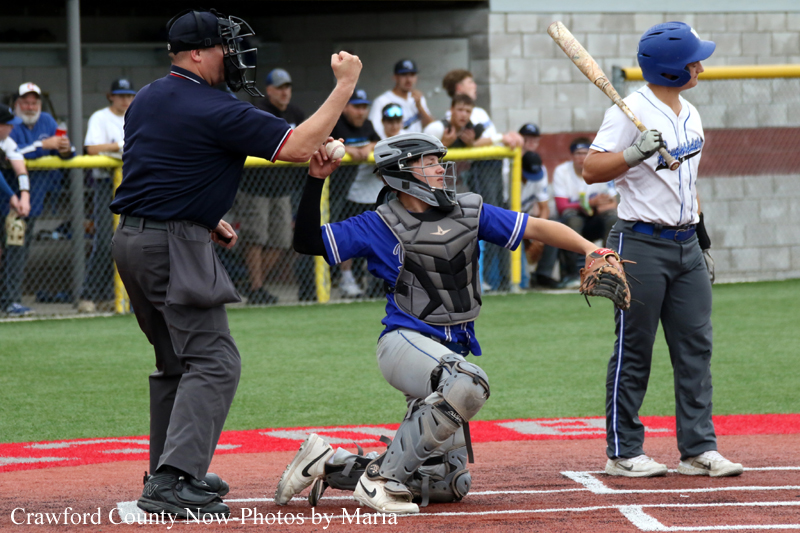 Baseball scene at home plate: catcher in blue and gray gear kneels as an umpire signals, while the batter waits in a white and blue uniform.