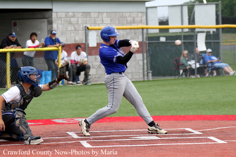 Baseball batter in a blue uniform swings at a pitch, with the catcher in position behind home plate and spectators in the background.